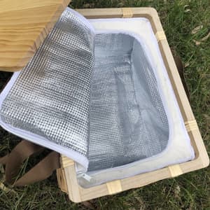 A wicker picnic cooler basket in natural wood with a silver insulated interior and an open lid, resting on grass.