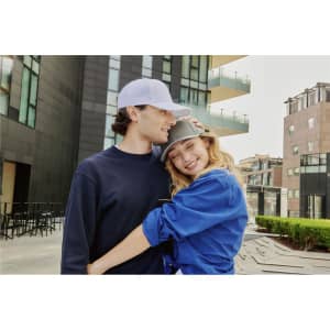 A couple is embracing outdoors, wearing a white cap and a grey cap, with modern buildings in the background.