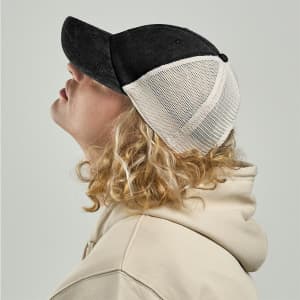 A black and white trucker cap displayed on a person with curly hair, wearing a beige hoodie, facing sideways.