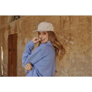 A smiling woman wearing a cream-coloured flat peak cap and a blue sweatshirt, set against a rustic wall background.