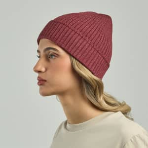 A maroon ribbed beanie worn by a model with wavy hair, facing to the side. The product has a logo.