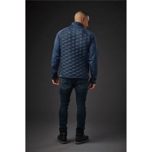 A man wears a navy blue thermal shell jacket with a quilted back, paired with dark jeans. The jacket has a logo.