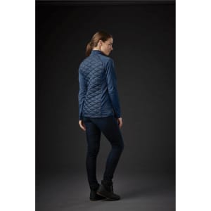 A woman wearing a blue quilted thermal shell jacket, viewed from the back, with dark jeans and footwear.