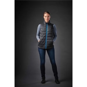 A woman is wearing a black thermal vest with a blue zip, paired with jeans and standing against a dark background.