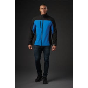 A man wearing a blue and black softshell jacket, standing against a dark background. The jacket has a zip and a logo.