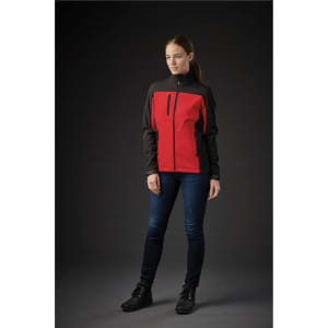 A woman is wearing a women's softshell jacket in red and black, with a zippered pocket, set against a dark background.