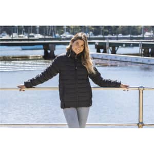 A woman wearing a black puffer jacket stands by the water, with a marina and bridge in the background.