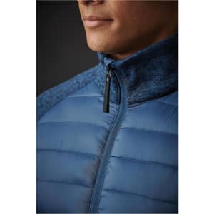 A close-up of a men's hybrid jacket featuring a blue quilted design and a high collar. The jacket has a zipper and a logo.