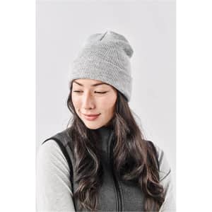 A grey knit beanie worn by a woman with long, wavy hair, paired with a dark vest. It features a logo.