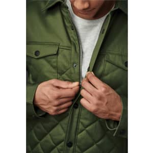 A man in a green quilted jacket adjusts the zipper, revealing a grey top underneath. The jacket features snap buttons and a logo.