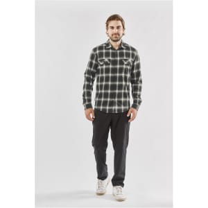 A man stands wearing a green and black checked button-up shirt with two chest pockets and black trousers.