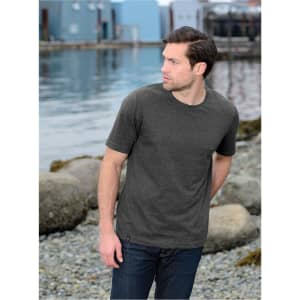 A man stands by a body of water wearing a dark grey short-sleeve t-shirt and jeans, with a rocky shoreline in the background.