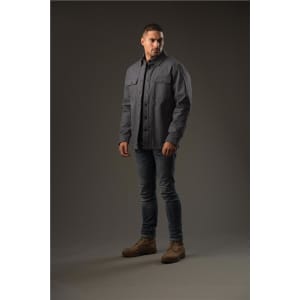 A man stands wearing a grey Men's Tradesmith Jacket, styled with fitted jeans and tan boots. The background is neutral.