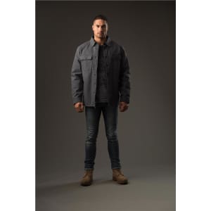 A man stands wearing a grey Men's Tradesmith Jacket over a grey top, paired with jeans and tan boots.