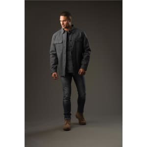 A man wearing a dark grey Tradesmith jacket over a shirt, paired with jeans and tan boots, standing against a neutral background.