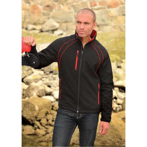 A man wearing a black softshell jacket with red accents, standing outdoors by rocky terrain. The jacket has a logo.