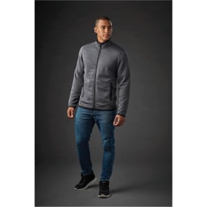A man wearing a grey Men's Andorra Jacket, paired with blue jeans and black shoes, standing against a dark background.