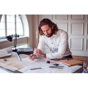 A man with long hair wearing a white button-up shirt, sitting at a desk with architectural plans and tools.