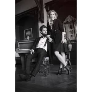 A woman in a black dress and high-heeled shoes stands next to a man in formal attire by a piano in a stylish room.