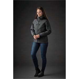 A woman stands in a black Women's Epsilon System Jacket, paired with blue jeans, against a dark background. The jacket features a logo.