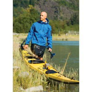 A man in a blue waterproof jacket beside a yellow kayak, with greenery and water in the background.