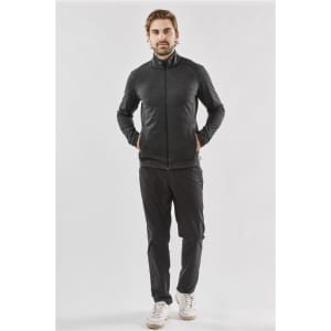 A man wearing a black performance jacket with a zip front and side pockets, paired with dark trousers and sneakers.