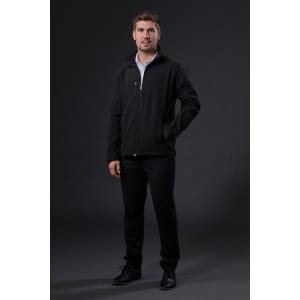 A man stands in a black softshell jacket, paired with black pants and shoes, against a dark background.