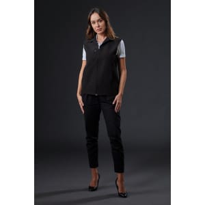 A black softshell vest is worn by a woman styled with sleek trousers and heels against a dark background. It features a logo.