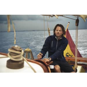 A man in a navy softshell jacket sits on a boat, with a stormy sea and a flag in the background.