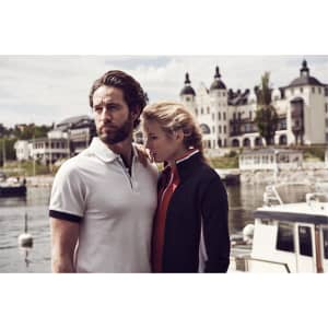 A man in a white polo shirt with black accents stands next to a woman in a dark jacket, near a waterfront.