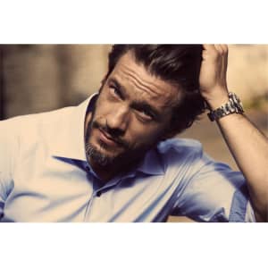 A stylish men's button-up shirt in light blue, featuring a logo, worn by a man with a beard.