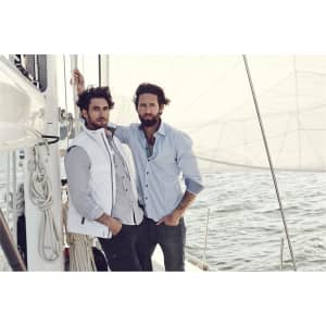 Two men posing on a sailboat, one wearing a light blue button-up shirt and the other in a white vest over a checked shirt.