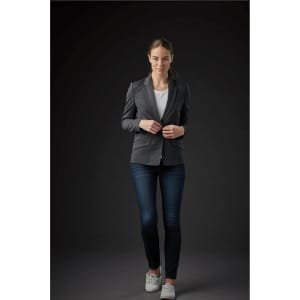 A woman in a grey knit blazer, white top, and jeans, standing against a dark background. The blazer has a logo.