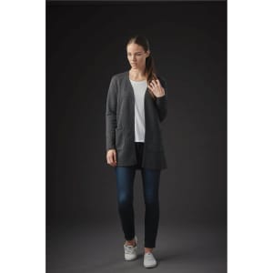 A woman wearing a grey quilted cardigan over a white top and jeans stands against a dark background.