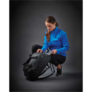 A woman in a bright blue softshell jacket is opening a black duffel bag. She is crouched down, focused on the bag.