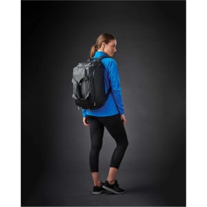 A woman wearing a blue lightweight softshell jacket, black leggings, and a black backpack, standing against a dark background.