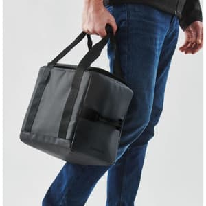 A cooler bag in black with sturdy handles, being carried by a person wearing jeans. Features a logo.