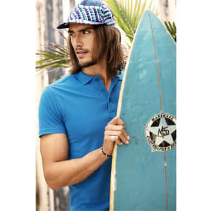 A man in a blue polo shirt holds a surfboard. He wears a patterned cap and stands near palm leaves.