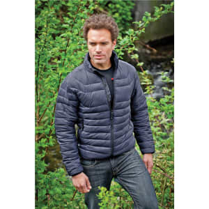 A man wearing a dark navy puffer jacket stands amidst lush greenery, with a creek visible in the background.