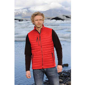 A man stands by a frozen lake wearing a red thermal vest over a black long-sleeve shirt, with icy landscapes in the background.