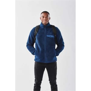 A man wearing a navy blue fleece jacket with a backpack, standing against a plain background.