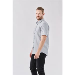 A light grey button-up shirt with short sleeves and a logo. The model is wearing black jeans.