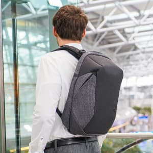 A person wearing a dark grey Swissdigital Cosmo 3.0 Backpack stands near a glass wall in a modern building.