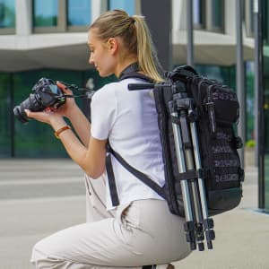 A woman with a black Swissdigital Berg XL Backpack crouches, holding a camera. The backpack has a tripod attached.