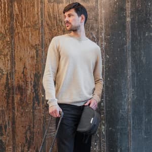 A man holding a black Swissdigital Arosa wash bag, wearing a beige sweater and standing against a textured wall.