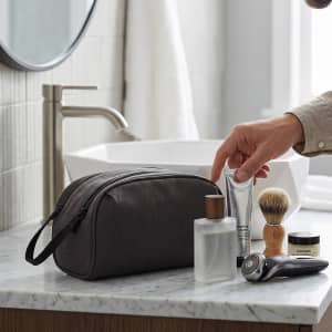 A grey wash bag alongside grooming products, including a bottle, tube, shaving brush, and razor on a marble countertop.