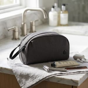 A grey toiletry bag sits on a marble countertop, accompanied by grooming products and a towel. It features branding.