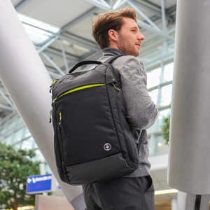 A black backpack with green accents is worn by a man in an indoor setting. The bag features multiple pockets and a logo.