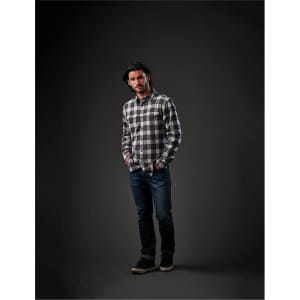 A man stands in a dark setting wearing a black and white checked snap front shirt and blue jeans.