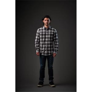 A man wearing a grey and black checkered snap front shirt, paired with dark jeans and shoes, against a dark background.